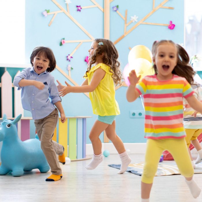 Drei Kinder, die fröhlich spielen und lachen in heller Frühlingskleidung in einem Raum mit der Hauptfarbe Hellblau.