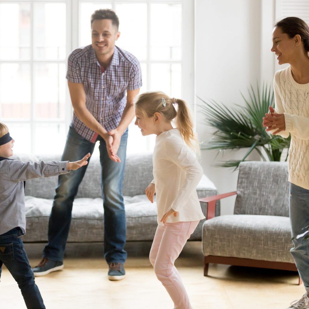 Blindfolded cute little boy playing hide and seek at home, parents and kids laughing spending time together enjoying game on weekend, happy family of four having fun leisure activity in living room